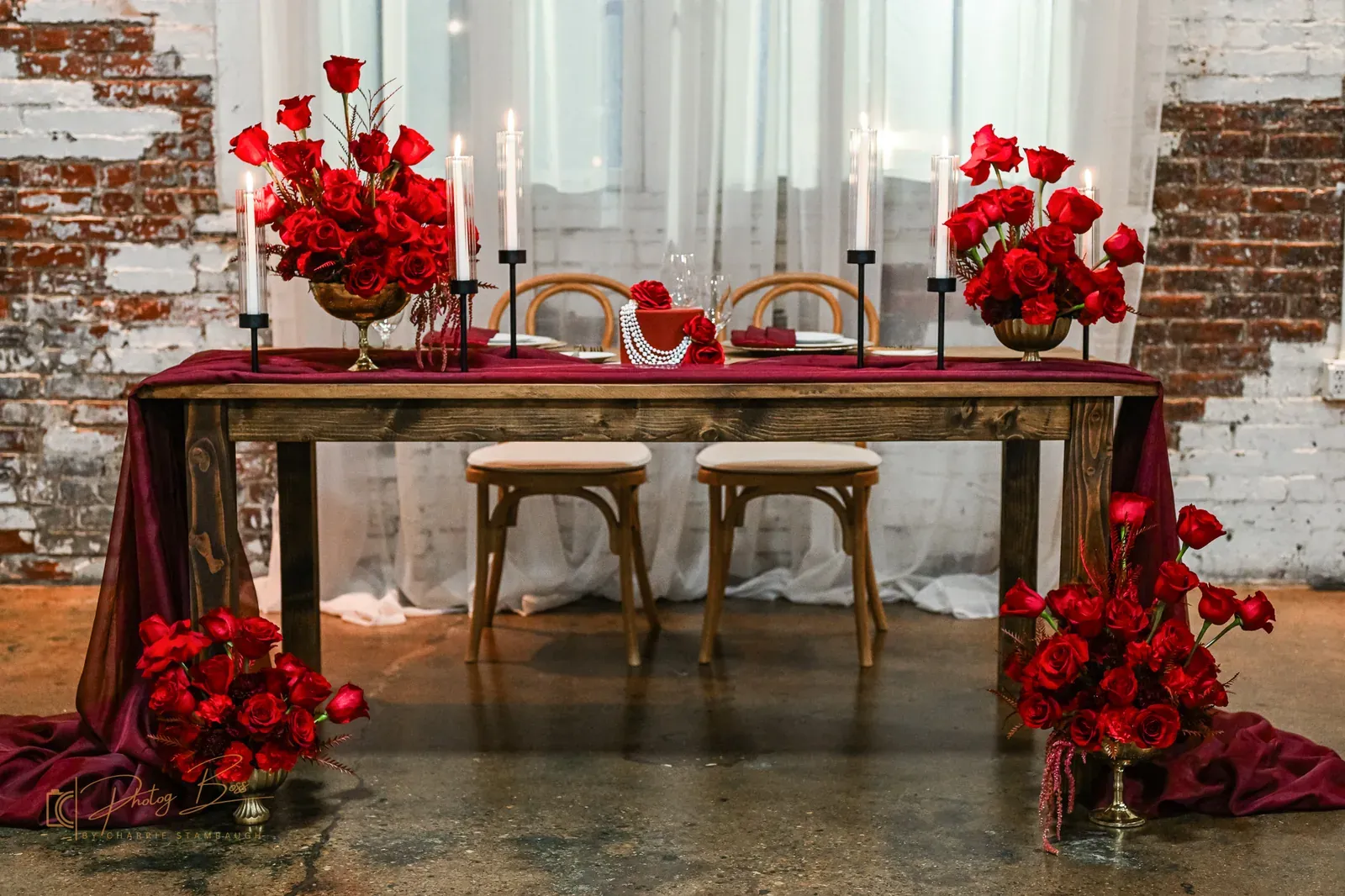 Sweetheart table at 24 Shelby with bold red rose arrangements, deep red runner, gold candle holders, and a wood farm table against original 1898 exposed brick, illustrating the 2026 Indianapolis wedding trend of bold color palettes