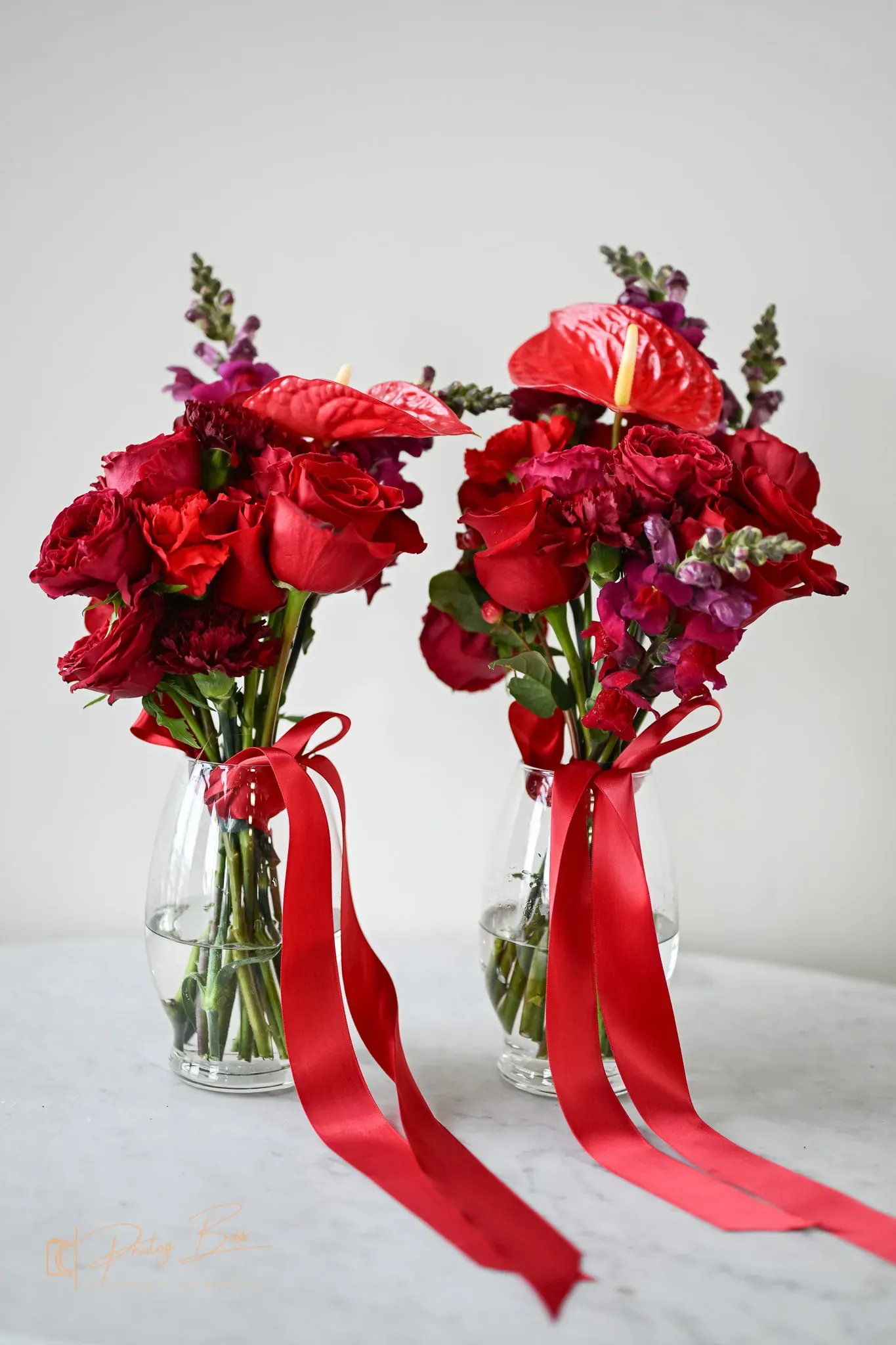 Two matching red rose and anthurium bouquets in glass vases tied with red satin ribbons. The bold-color floral direction trending across Indianapolis bridal showers and weddings in 2026