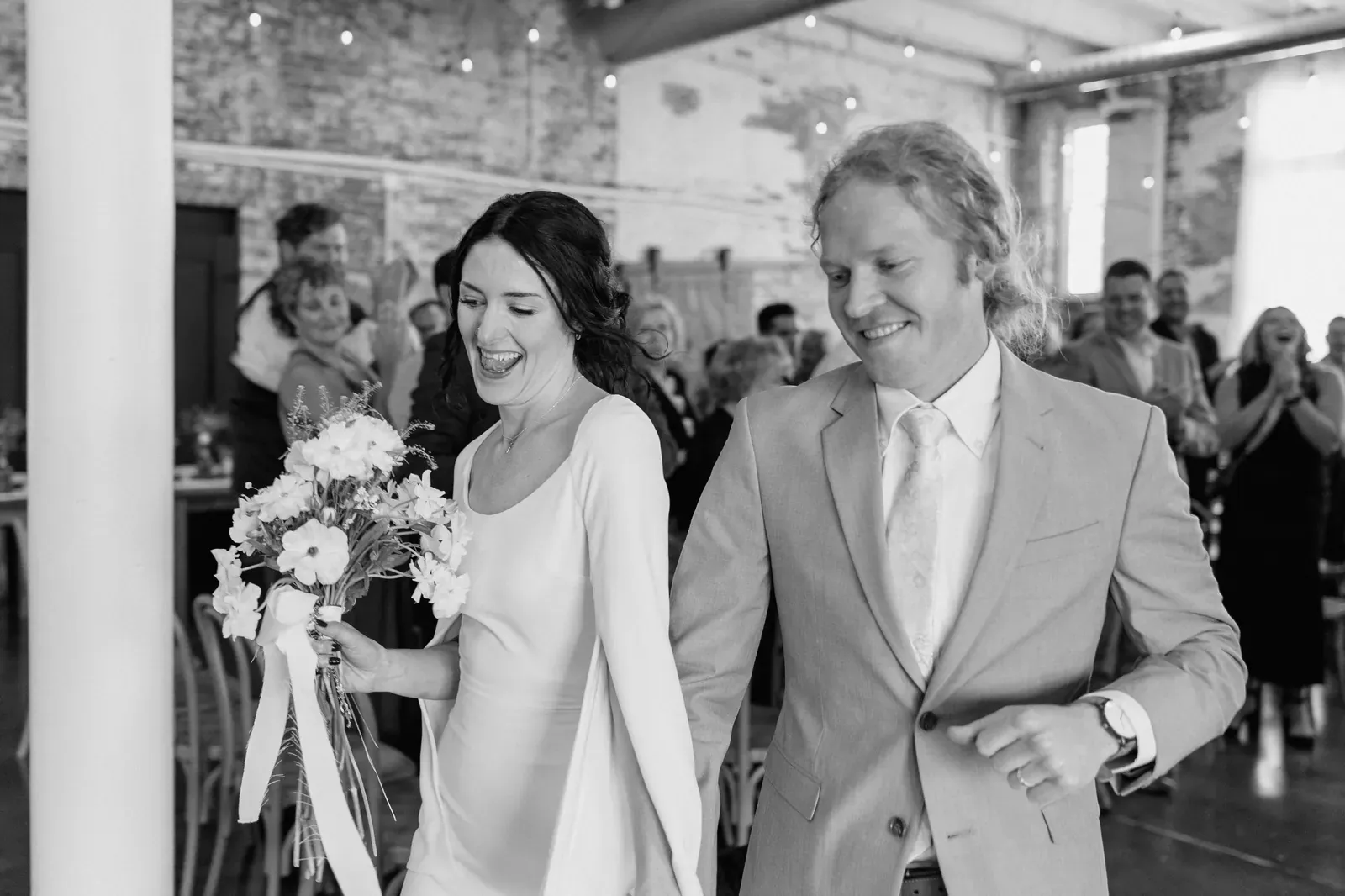 Couple making their grand entrance into the reception as cheering Indianapolis wedding guests fill the floor under Edison string lights