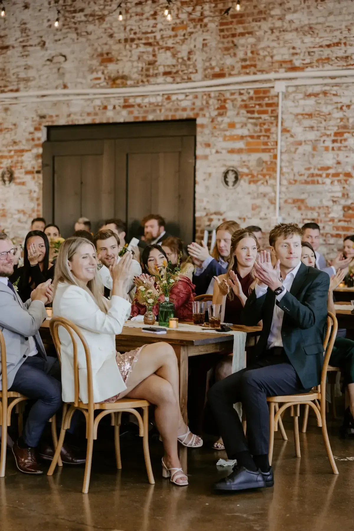 Guests at a long table at 24 Shelby laughing and applauding a live performance under the Edison Room's brick walls