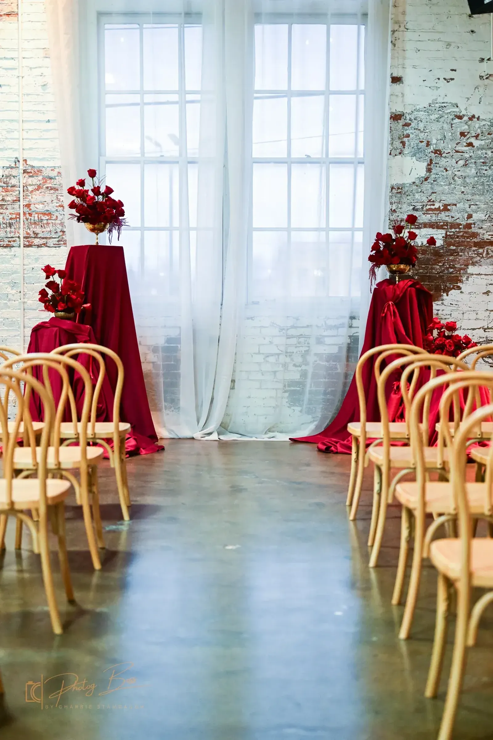 Indoor ceremony at 24 Shelby for an Indianapolis wedding in 2026, with rows of light wood chairs facing an altar of bold red drapery and oversized red and gold floral arrangements against original 1898 brick walls.
