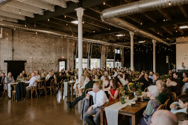 The Edison Room at 24 Shelby in downtown Indianapolis set for a corporate reception with round tables, ambient uplighting, and Edison string lights overhead against the original 1898 exposed brick walls.