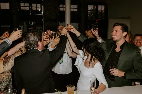 Guests raise cocktails in a group toast at an evening engagement party in a downtown Indianapolis venue, under Edison string lights against exposed brick and tall industrial windows.
