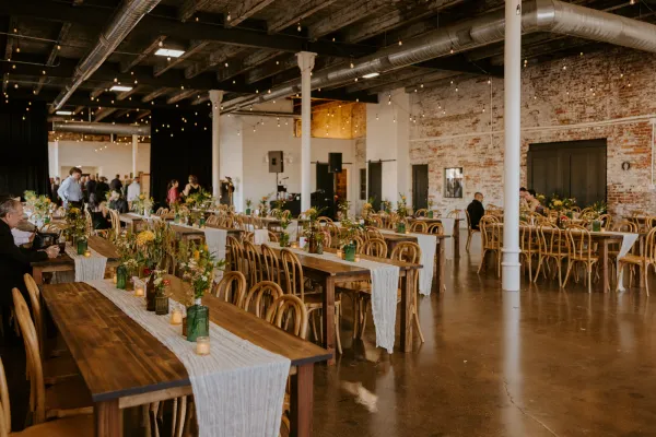 The Edison Room at 24 Shelby set up for an Indianapolis wedding reception with long tables, ghost chairs, Edison string lights, original 1898 exposed brick walls, and wood-beam ceilings. The downtown Indianapolis venue character that does most of the decorating for couples on a budget.