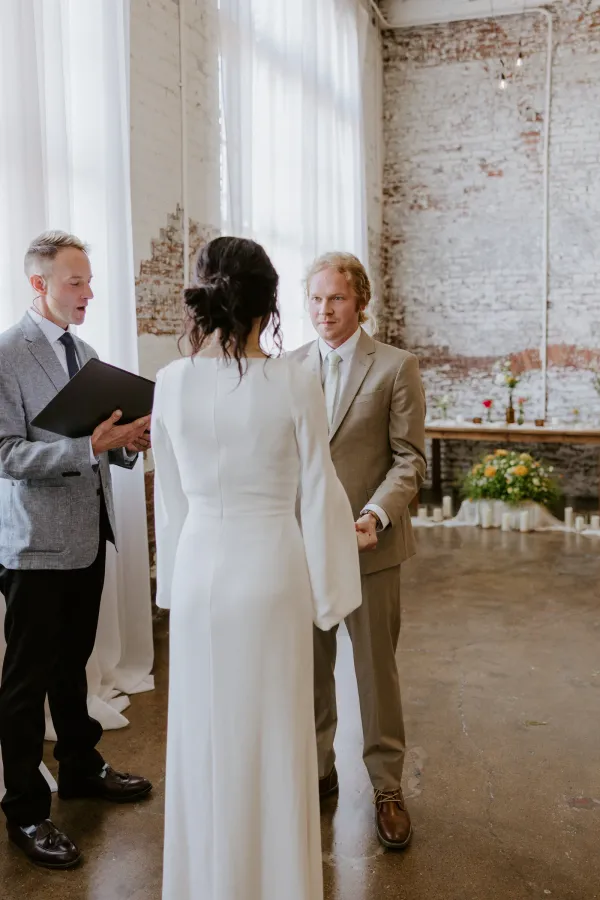 Intimate wedding ceremony at 24 Shelby in downtown Indianapolis with the bride and officiant standing in a softly lit historic space, exposed brick walls and Edison string lights overhead, set up for a small celebration of fewer than 75 guests.