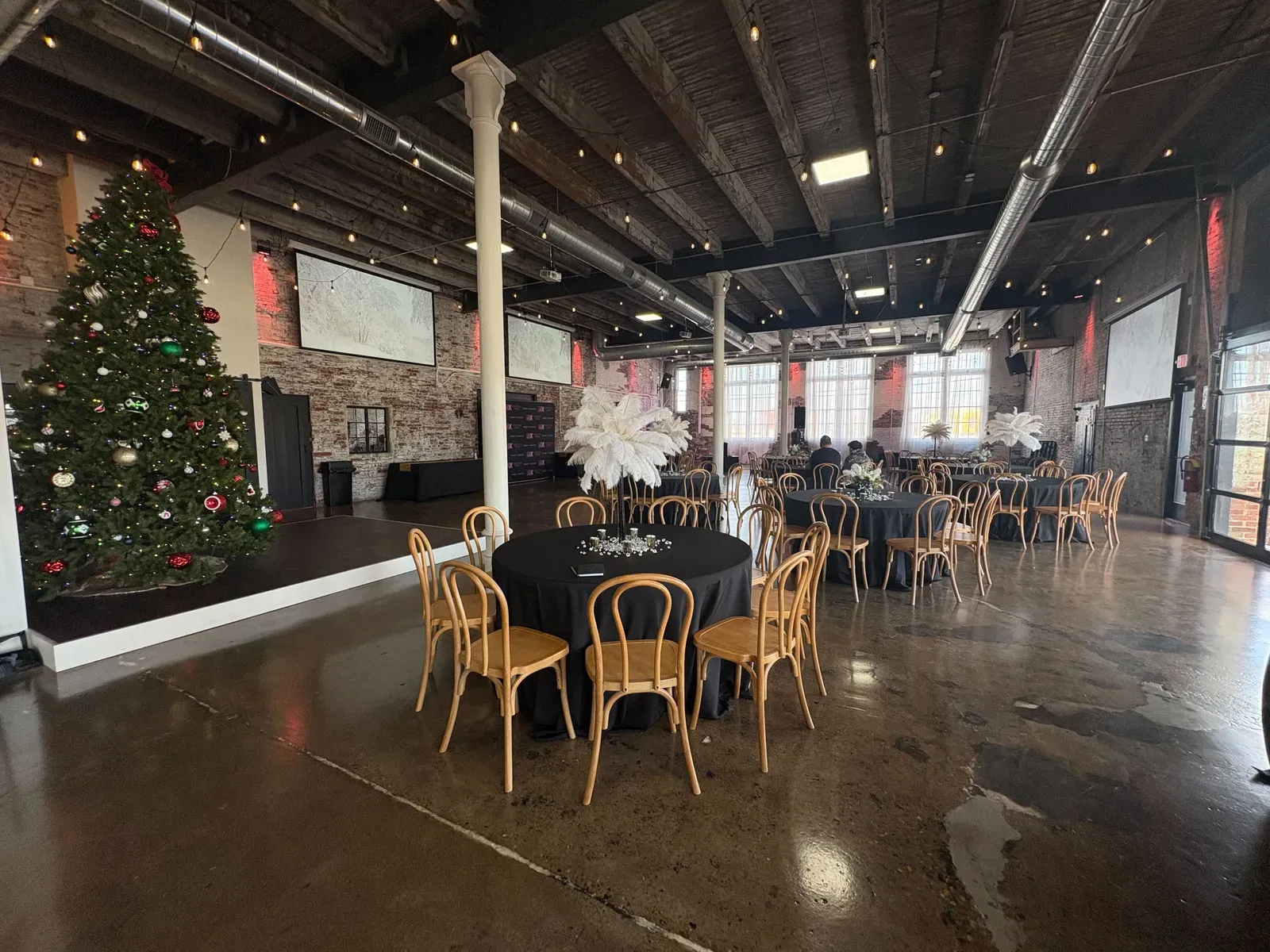 A corporate holiday party setup at 24 Shelby in downtown Indianapolis with black-linen round tables, gold chairs, wall projection screens, and a festive tree