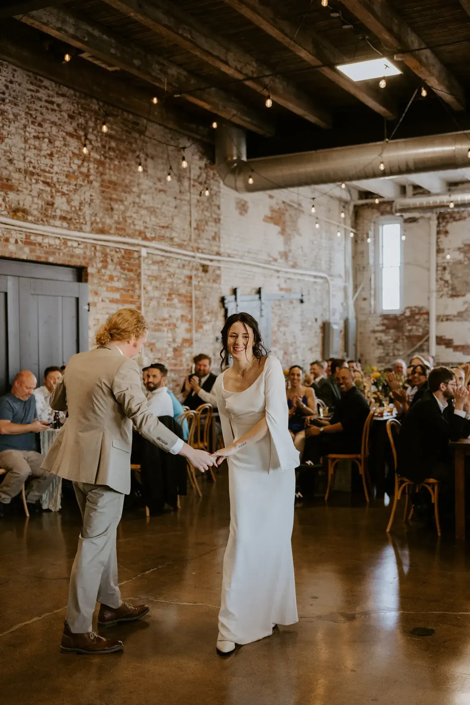 Reception in full swing inside an affordable historic Indianapolis wedding venue with exposed brick walls, Edison string lights, and high wood-beam ceilings.