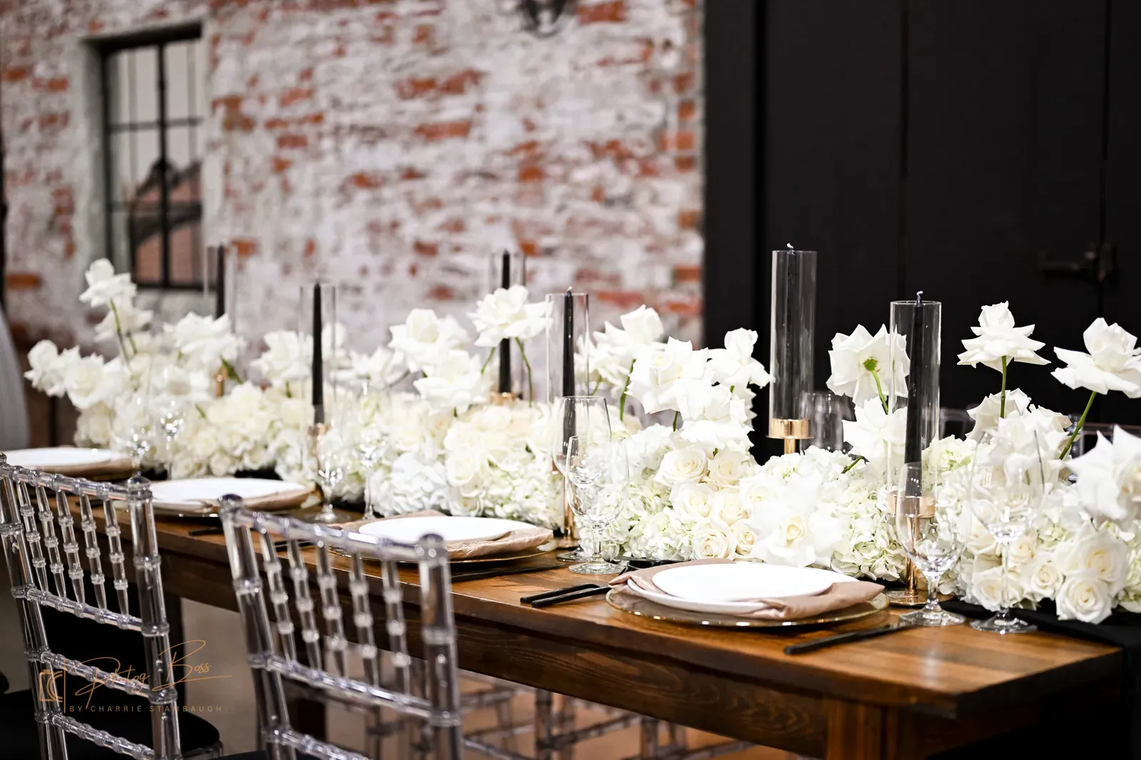 Long wood reception table with white-rose runner, gold chargers, ghost chairs, and exposed brick at 24 Shelby for an Indianapolis wedding
