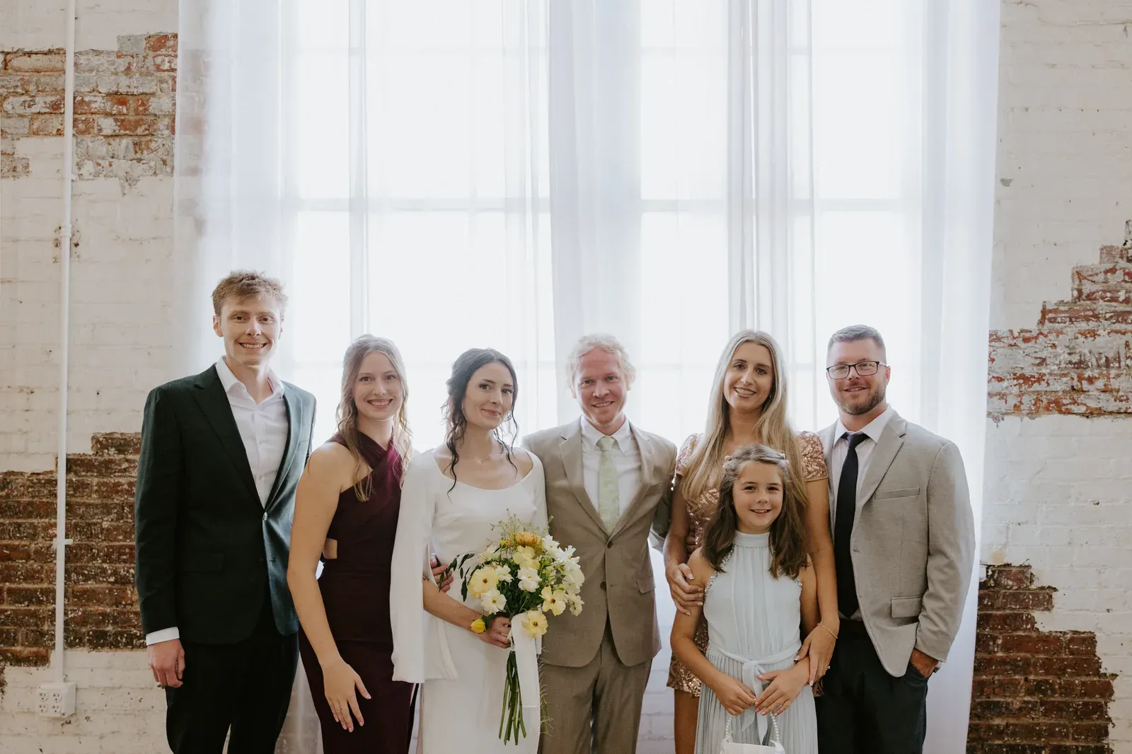 A small wedding party gathering inside an industrial-chic Indianapolis venue with exposed brick walls, large windows, and warm Edison lighting overhead